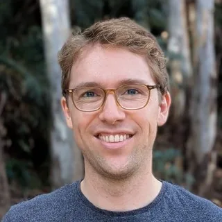 Portrait of Simon de Waal, smiling, wearing glasses and a blue-grey sweater, with eucalyptus trees behind him